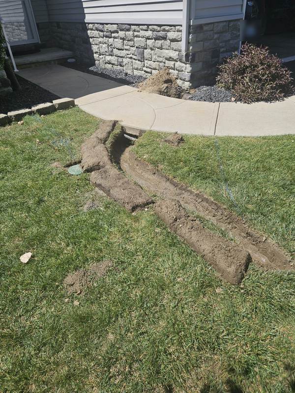 Open trench cut through a front lawn toward the downspout, with strips of sod rolled back alongside it
